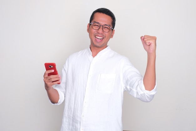 Photo adult asian man cheering happy while holding a mobile phone