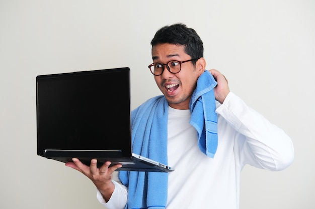 Photo adult asian man doing online meeting using laptop while still wearing sleeping clothes and towel around his neck