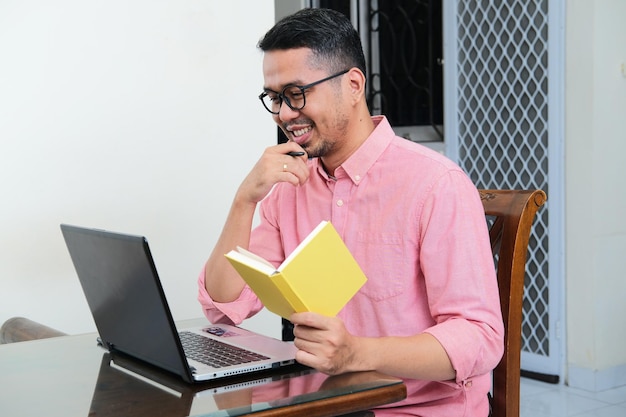 Uomo asiatico adulto che tiene un libro mentre guarda al suo computer portatile con l'espressione felice