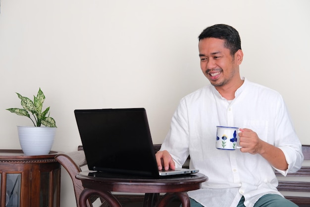 Adult Asian man holding a glass of drink while checking email on his laptop