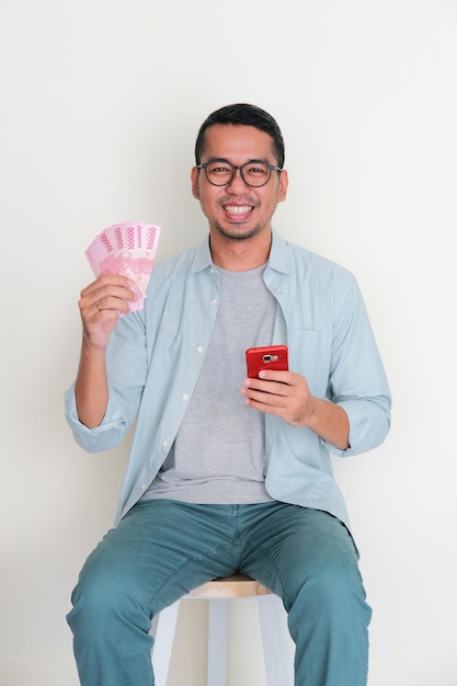 Adult Asian man holding handphone and paper money sitting in a bench