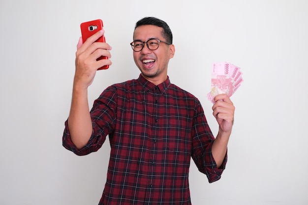 Взрослый азиатский мужчина держит деньги и показывает счастливое выражение лица, глядя на свой телефон