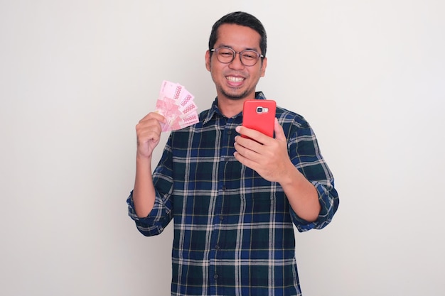 Photo adult asian man holding money smiling happy while looking to his phone
