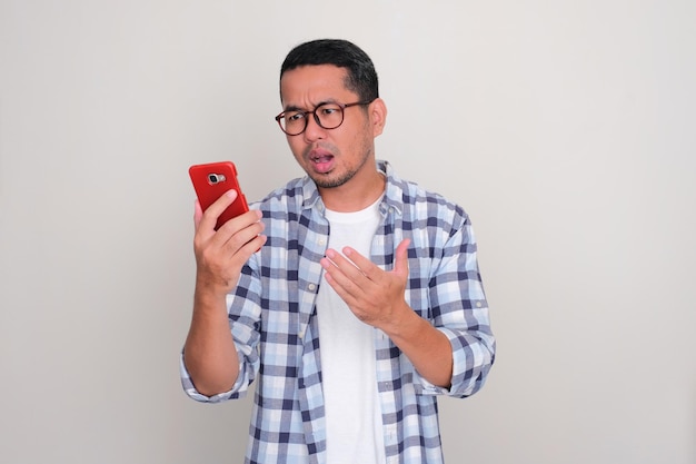 Взрослый азиат смотрит на свой мобильный телефон с разочарованным выражением лица