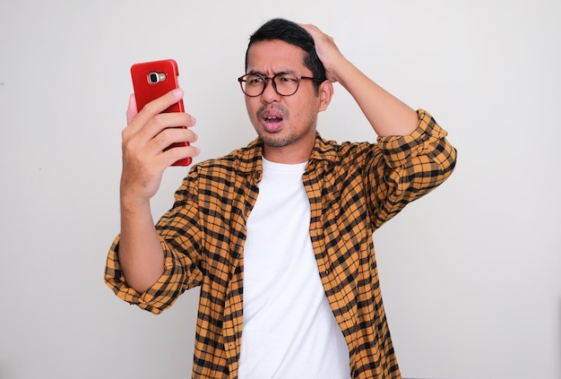 Adult Asian man looking to mobile phone that he hold with confused expression