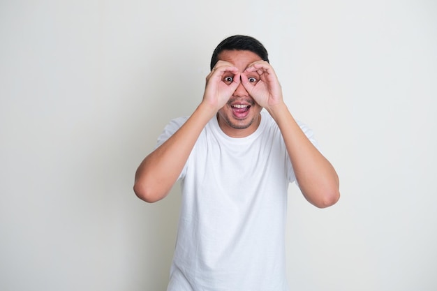 Adult Asian man making binoculars shape with his hands and looking through it with excited expression