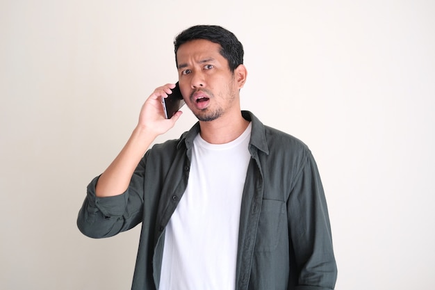 Photo adult asian man showing confuse face expression while answering a call using mobile phone