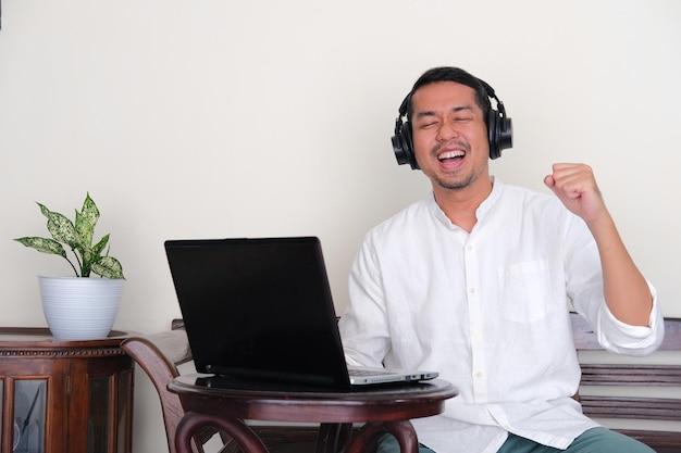 Photo adult asian man showing happiness when listening music and sitting in front of laptop