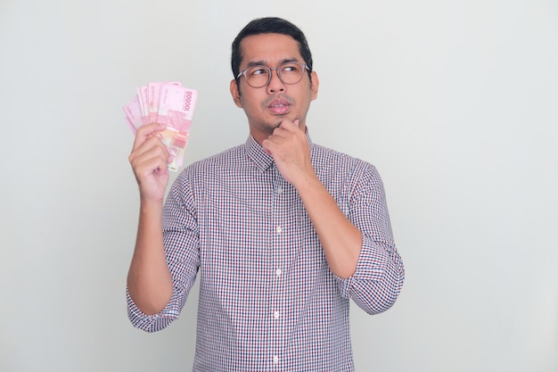 Adult Asian man showing thinking expression while holding money