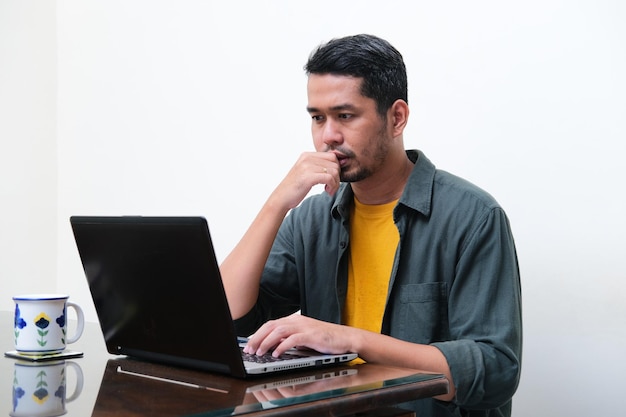 Adult Asian man showing thinking gesture in front of his laptop