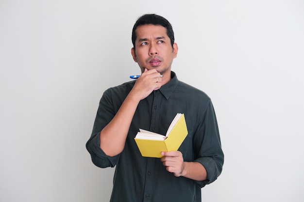 Photo adult asian man showing thinking gesture while holding pen and note book