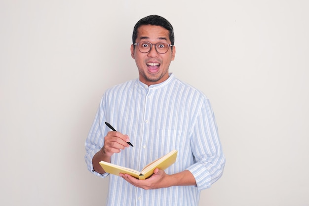 Photo adult asian man showing wow face expression when holding a book and pen