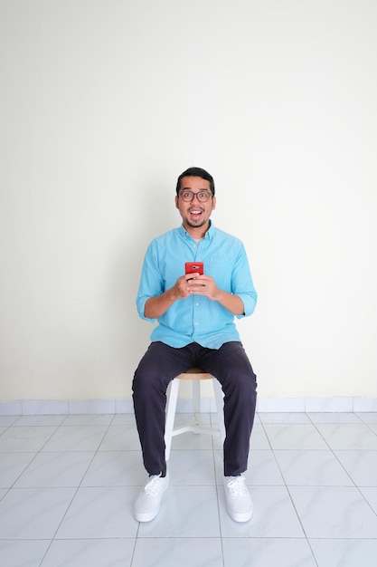 Взрослый азиатский мужчина сидит на скамейке и держит мобильный телефон с возбужденным выражением лица