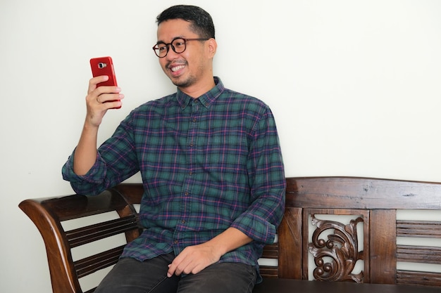 Photo adult asian man sitting in a bench showing happy expression while looking to his mobile phone