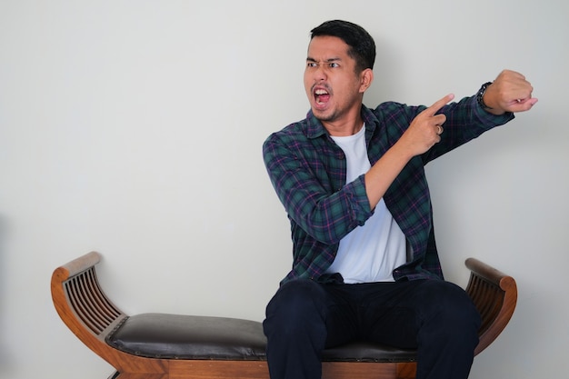 Adult Asian man sitting in a chair and showing angry face expression while pointing to his arm watch