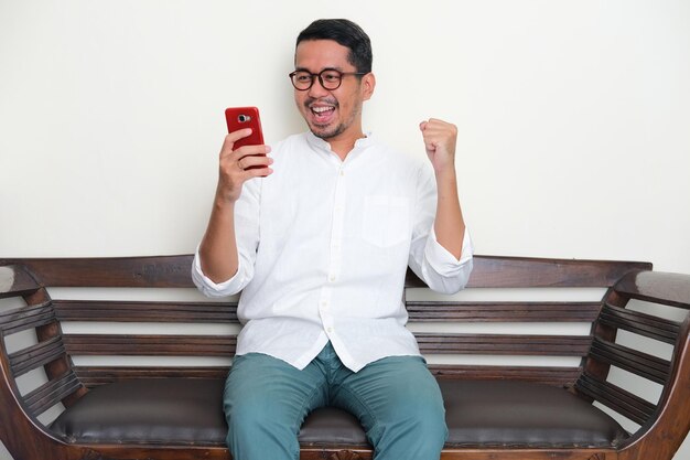 Photo adult asian man sitting in a couch clenched fist showing excited when looking to mobile phone