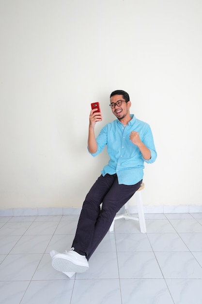 Adult Asian man sitting relax in a bench looking to his handphone with excited