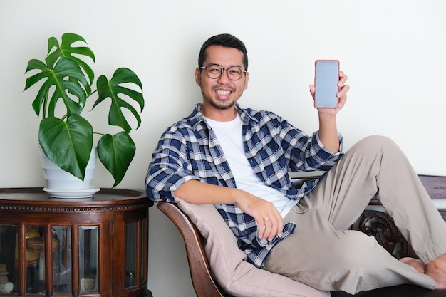 Adult Asian man sitting relax in a couch while showing blank mobile phone screen