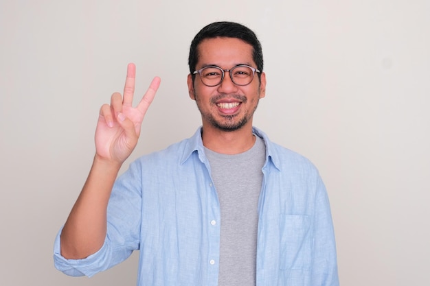 Взрослый азиатский мужчина улыбается в камеру и делает знак двумя пальцами