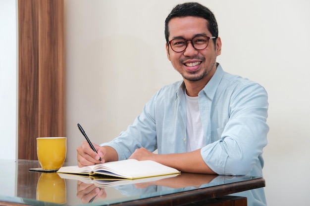 Adult Asian man smiling at camera while writing in a book