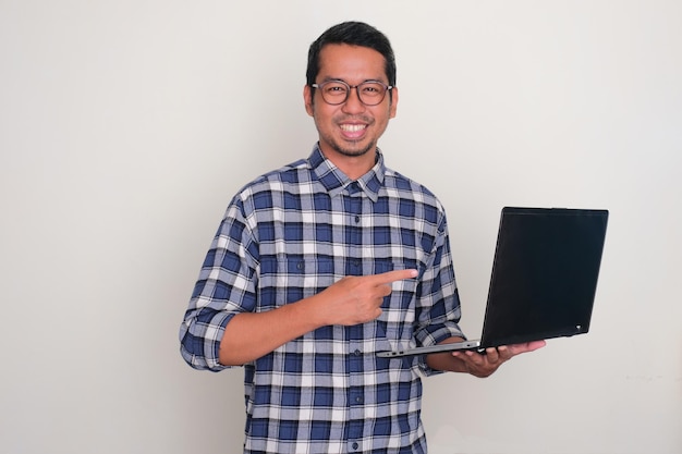 Photo adult asian man smiling confident and pointing to a laptop that he hold