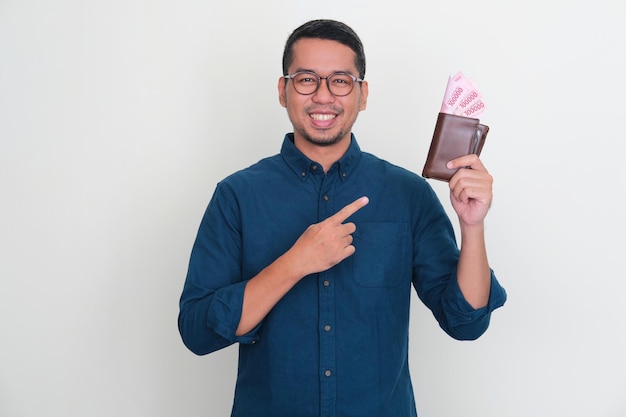 Adult Asian man smiling confident while pointing to wallet full of money that he hold