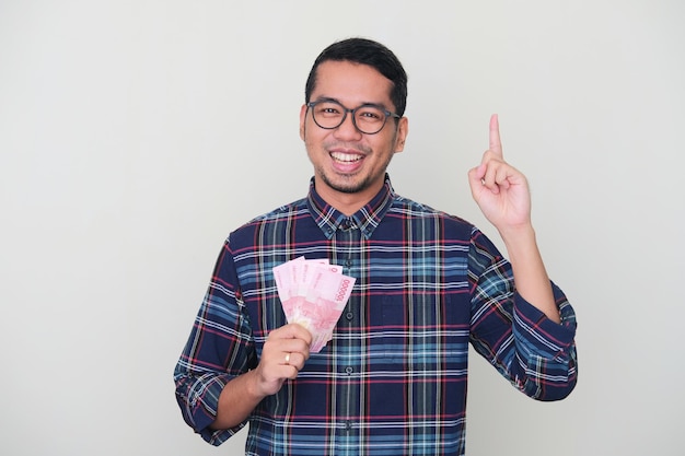 Взрослый азиат счастливо улыбается и показывает пальцем вверх, держа бумажные деньги Индонезии