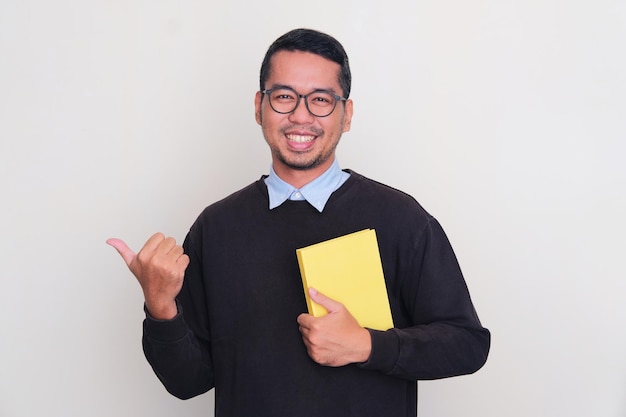 Photo adult asian man smiling happy and pointing to the right side while holding a book