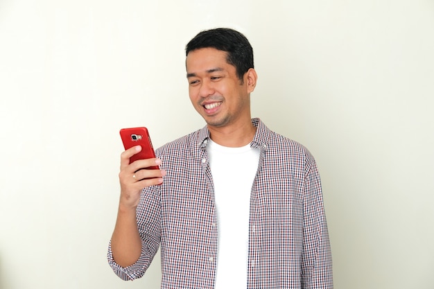 Взрослый азиатский мужчина улыбается счастливым, глядя на экран своего мобильного телефона