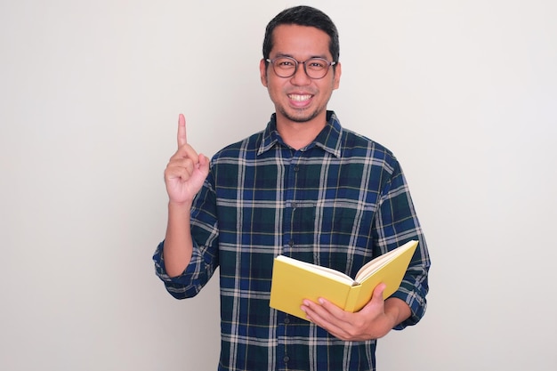 Photo adult asian man smiling and pointing finger up while holding a book