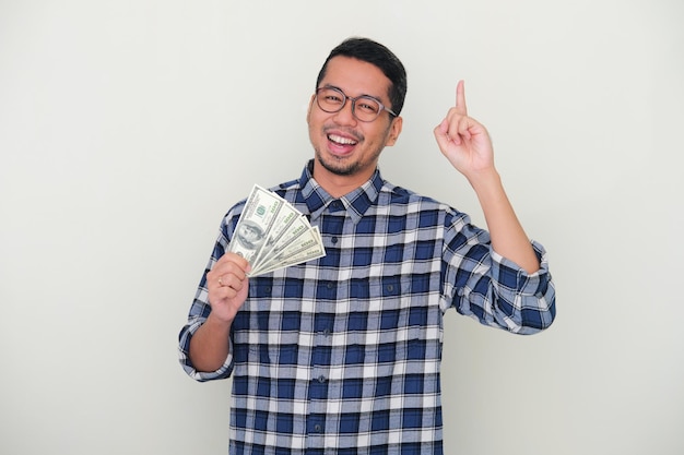 Adult Asian man smiling and pointing finger up while holding united states dollar paper money