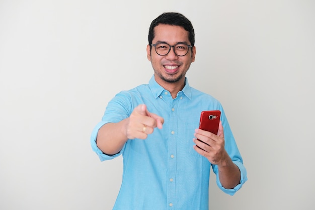 Взрослый азиатский мужчина улыбается и показывает пальцем вперед, держа мобильный телефон