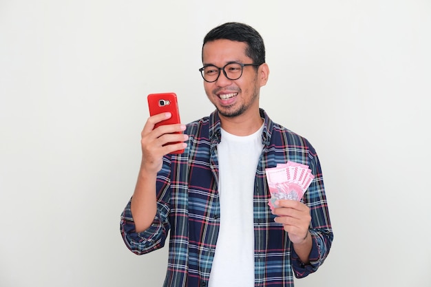 Adult Asian man smiling when looking to his handphone while holding money