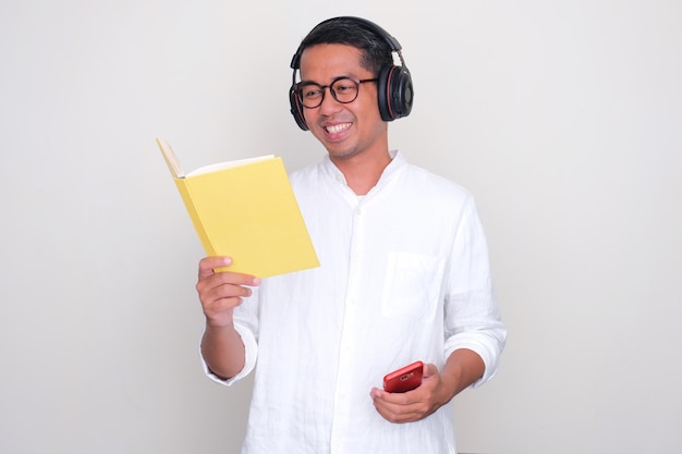 Photo adult asian man smiling when reading a book and listening music using earphones