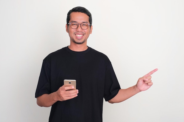 Взрослый азиатский мужчина улыбается, держа телефон, одной рукой указывая влево.