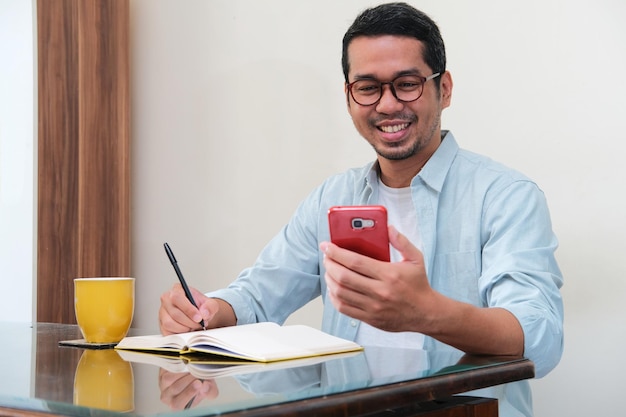 彼の携帯電話を見ながら笑みを浮かべて、本に書く大人のアジア人男性