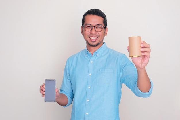 Photo adult asian man smiling while showing a canned product and holding blank mobile phone screen