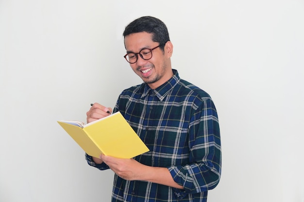 Photo adult asian man smiling while writing something on a book