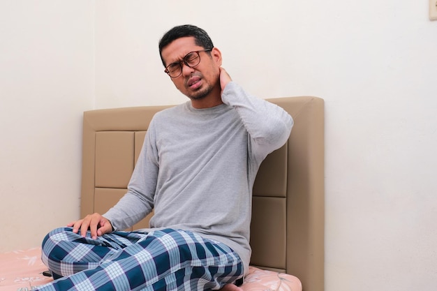 Adult Asian man suffering neck pain while sitting on his bed