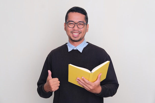 Adult Asian man wearing black sweater smiling and give thumb up while holding a book