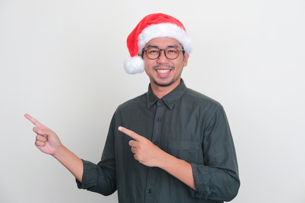 クリスマス帽子をかぶった大人のアジア人男性が、右側を指差しながら幸せそうに笑っている
