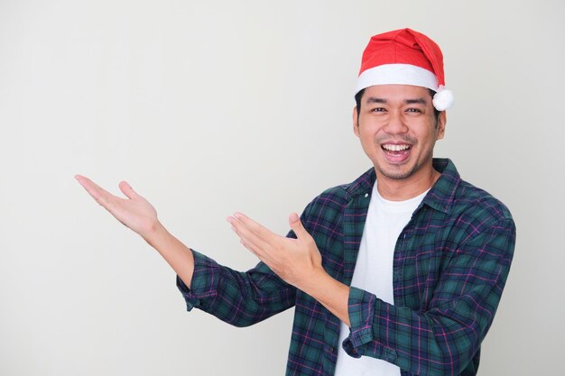 Adult Asian man wearing christmas hat smiling happy while showing beside