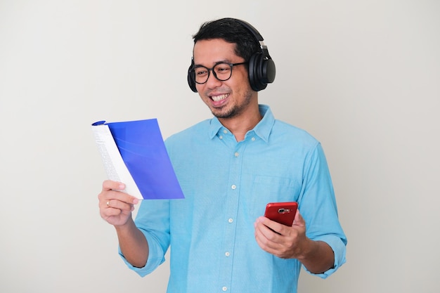 Uomo asiatico adulto che indossa le cuffie e tiene in mano il cellulare quando legge un libro