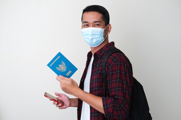 Uomo asiatico adulto che indossa una maschera medica protettiva con in mano un telefono e un passaporto del paese indonesiano