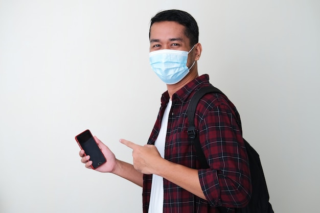 Photo adult asian man wearing protective medical mask holding handphone and pointing finger on it