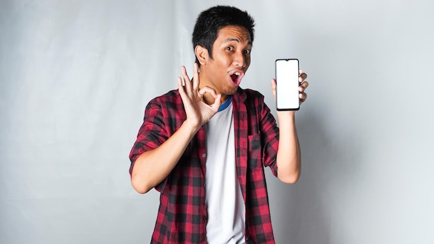 Фото Взрослый азиат в красной фланелевой рубашке улыбается и показывает большой палец вверх, держа мобильный телефон и жест рукой ок