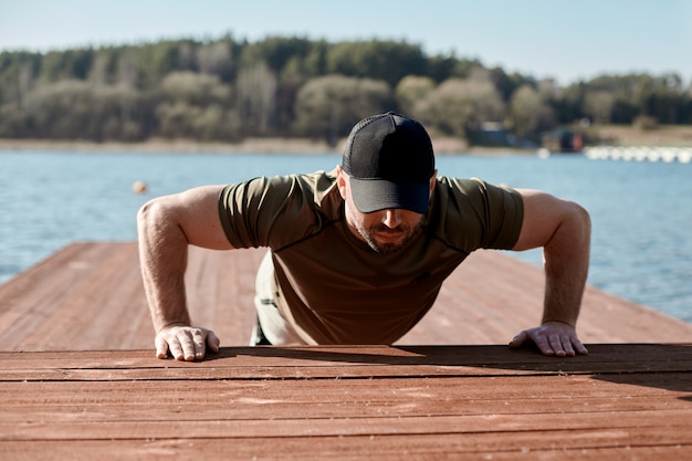 Foto un adulto uomo atletico esegue push-ups su un molo sul lago. un uomo è impegnato in sport all'aria aperta. sport, fitness, stile di vita