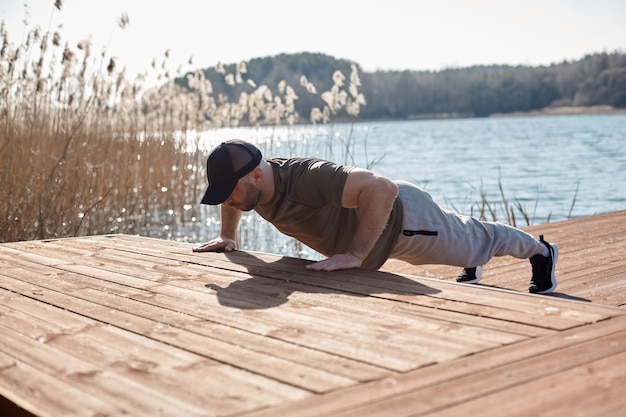 An adult athletic man performs push-UPS on a pier on the lake. A man is engaged in outdoor sports. Sports, fitness, lifestyle