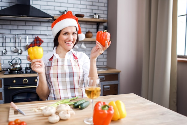 Adult attractive woman stand in kitchen and posing. Holding peppers in hands. Wear red festive hat. Celebrating Christmas or new year. Cooking alone.