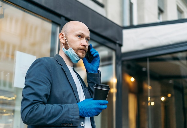 Adult bald bearded man in medical face mask and rubber gloves\
with paper cup of coffee to go with mobile. new normal, concept of\
coronavirus quarantine